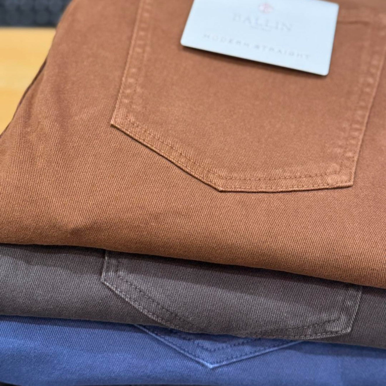 Stack of folded brown, blue, and gray pants with a brand tag on a wooden surface.